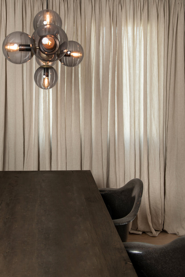 Dark wooden dining table set with chairs under a stylish lamp, framed by elegant beige semi-transparent linen drapes in the fabric breeze oatmeal by ann rees.
